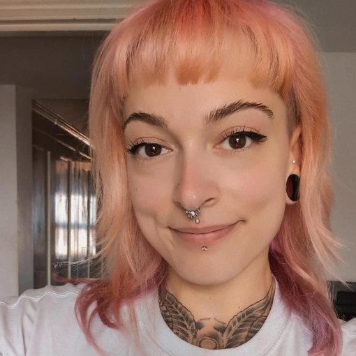 A person with light pink hair, septum and lip piercings, gauged ears, and visible chest tattoos smiles at the camera indoors. A person with light pink hair, septum and lip piercings, gauged ears, and visible chest tattoos smiles at the camera indoors.