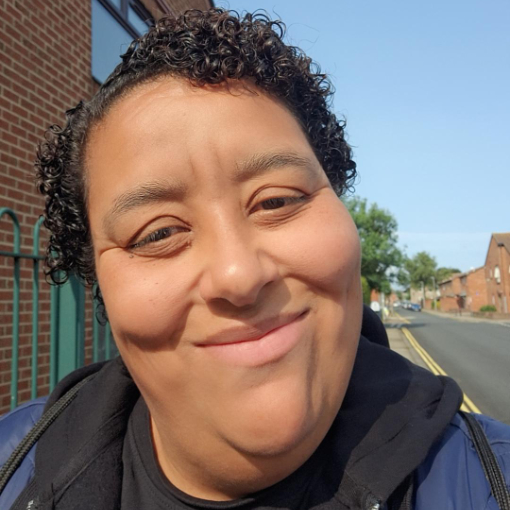 A person with short curly hair smiles at the camera while standing outside on a sunny street with brick buildings in the background. A person with short curly hair smiles at the camera while standing outside on a sunny street with brick buildings in the background.