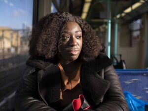 A person with curly hair and a serious expression sits on a train, looking out the window with sunlight illuminating their face. They are wearing a dark coat and holding items in their lap.