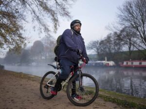 A person in a helmet and jacket stands beside a bicycle on a path next to a misty river, with trees and boats in the background.