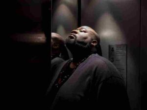 A man leans against the mirrored wall of an elevator, eyes closed and head tilted back, under dim lighting.