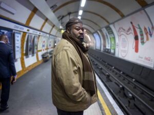 A man in a beige jacket and scarf stands on a subway platform, gazing slightly to his right. Other commuters and advertising posters are visible in the background.