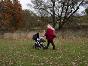 A person walks on grass pushing a baby stroller, surrounded by trees and autumn leaves.