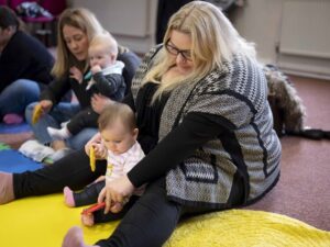 Adults sitting on a carpet with babies; one woman holds a baby in her lap, while another baby sits on the floor playing.