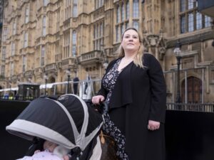 A woman stands outdoors in front of an ornate building, next to a stroller with a baby covered by a blanket.