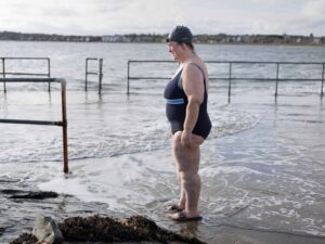 Person standing on a beach, wearing a swimsuit, swim cap, and goggles, looking out at the ocean. There are metal railings and foamy waves nearby, with a distant shoreline in the background.