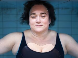 A person with short curly hair and a neutral expression is submerged in water, only their head and shoulders visible. They are wearing a black swimsuit against a tiled background.