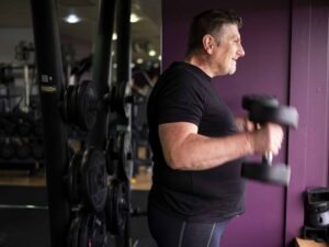 A man in a black t-shirt performs bicep curls with dumbbells in a gym, standing beside a rack of weights.
