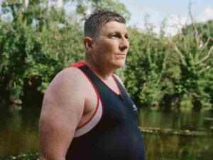 A man wearing a sleeveless shirt stands outdoors near a body of water, with trees and greenery in the background.