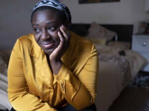 A person in a mustard-colored top, with a patterned headscarf, sits in a bedroom, resting their head on their hand and smiling slightly.