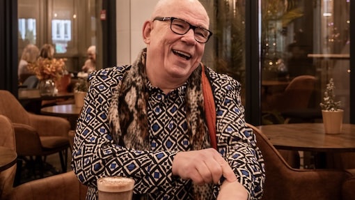 An elderly person wearing glasses and a patterned shirt is smiling while seated at a café table with a glass of coffee in front.