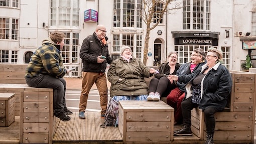 A group of six people sitting and conversing on outdoor wooden benches in an urban setting.
